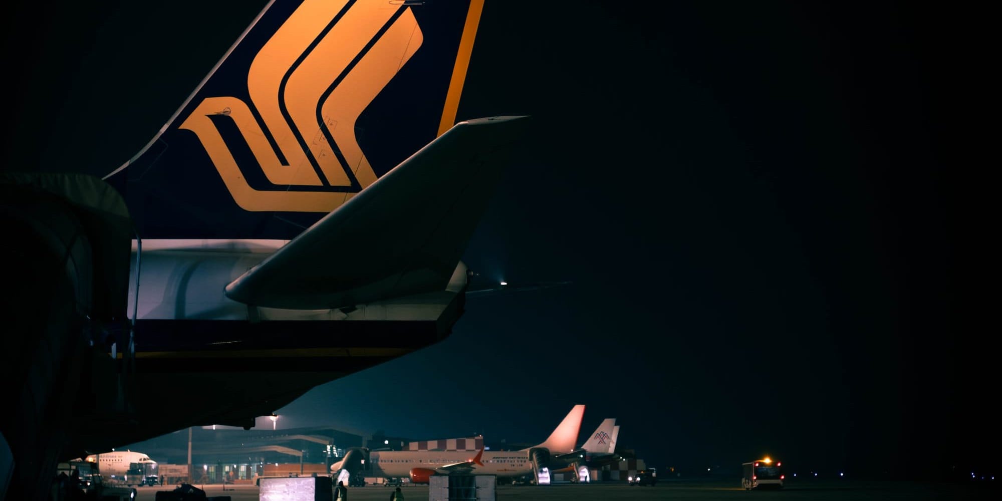The tail of a Singapore Airlines aircraft lit against a near-black night apron, ground crew and parked aircraft small in the distance