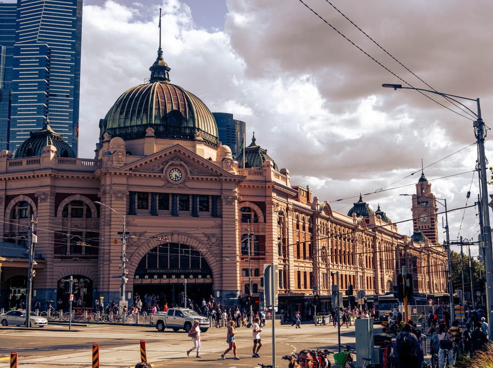 Sandstone railway station with a green copper dome, crowds crossing in front under a heavy afternoon sky